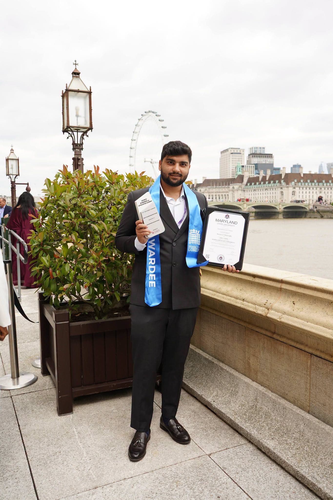 Mr Muhammad Faizan Khan holding his honorary doctorate certificate beside the River Thames and Palace of Westminster